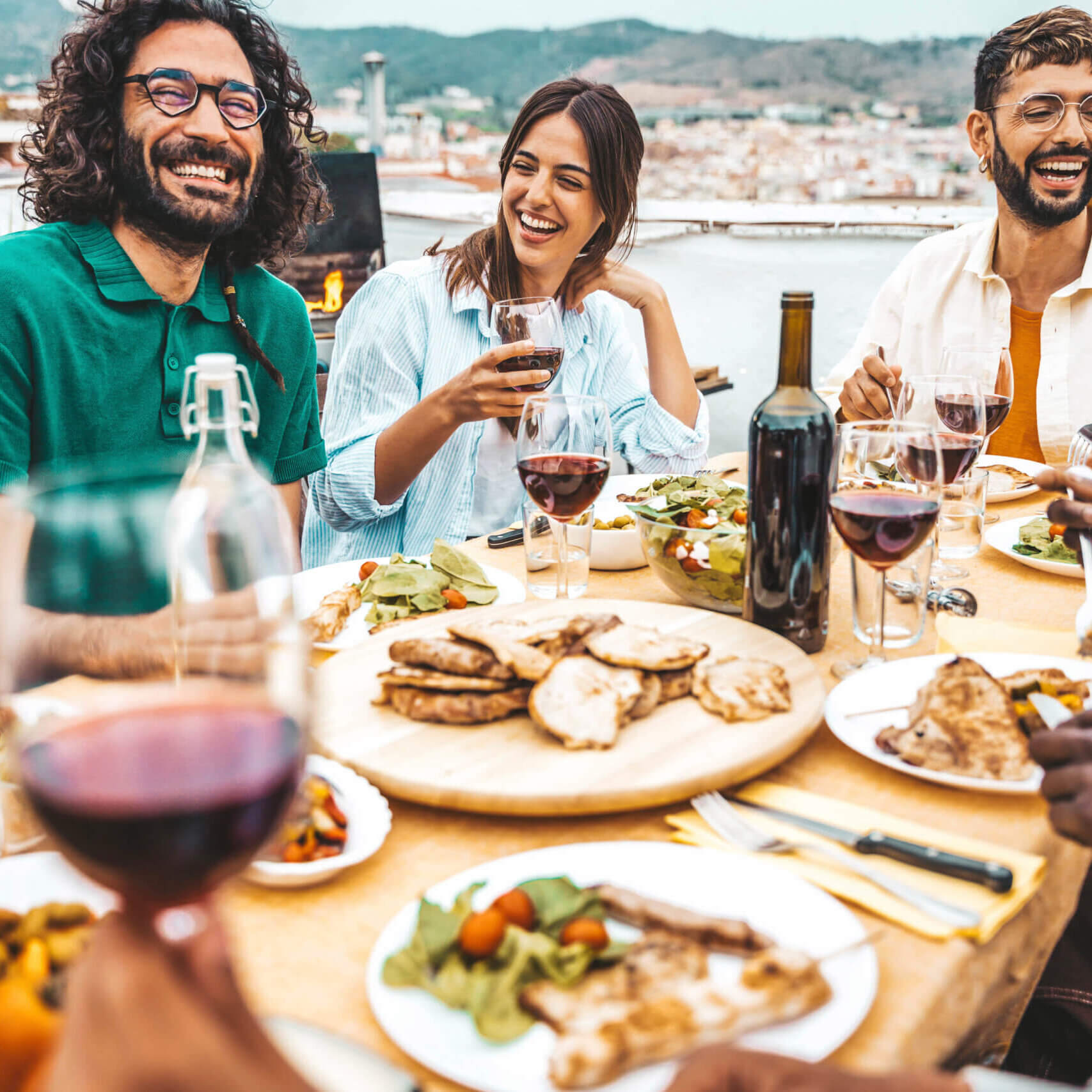 Multiethnic friends having fun at rooftop bbq dinner party - Group of young people diner together sitting at restaurant dining table - Cheerful multiracial teens eating food and drinking wine outside
